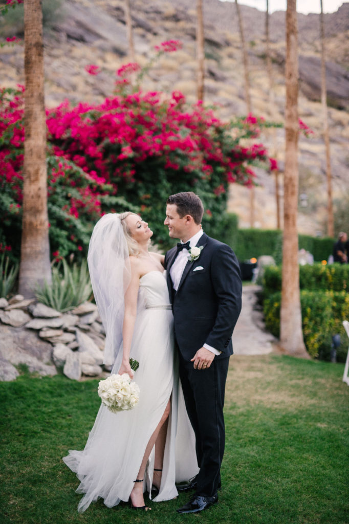 Casual portrait of bride and groom on their ceremony lawn, with bougainvillea behind them. 
